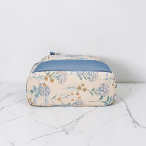 May include: A floral-patterned backpack with a light blue top panel. The bag features a cream-coloured base with a repeating pattern of blue and yellow flowers and green leaves. The bag is sitting on a white marble surface.