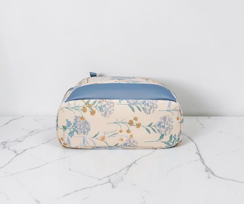 May include: A floral-patterned backpack with a light blue top panel. The bag features a cream-coloured base with a repeating pattern of blue and yellow flowers and green leaves. The bag is sitting on a white marble surface.