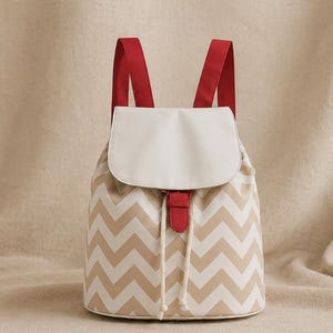 May include: A backpack featuring a beige and white chevron pattern on the main body, a solid white flap closure, red shoulder straps, and a red tab accent over a drawstring closure with two white cords hanging down the front of the bag.