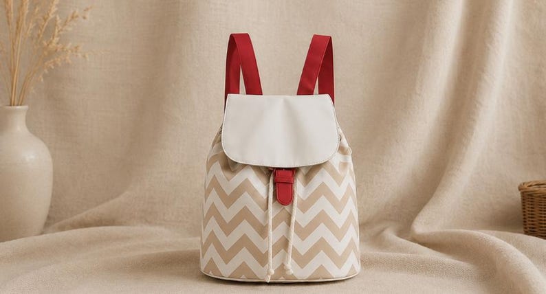 May include: A backpack featuring a beige and white chevron pattern on the main body, a solid white flap closure, red shoulder straps, and a red tab accent over a drawstring closure with two white cords hanging down the front of the bag.