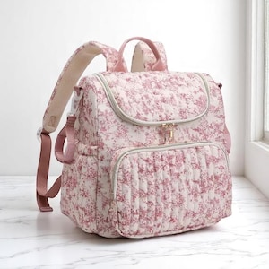 Puede incluir: Una mochila acolchada con un estampado floral en tonos rosa y blanco. La bolsa tiene un asa superior, correas ajustables para los hombros y m&uacute;ltiples compartimentos con cremallera. Los herrajes son dorados.