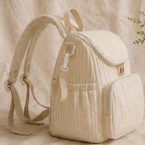 May include: A cream-coloured quilted backpack featuring a vertical ribbed texture, a top handle, adjustable padded shoulder straps, and a front zipped pocket. The bag includes gold-toned metal zip pulls and side elasticated pockets for storage.