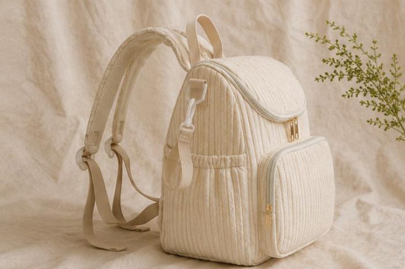 May include: A cream-coloured quilted backpack featuring a vertical ribbed texture, a top handle, adjustable padded shoulder straps, and a front zipped pocket. The bag includes gold-toned metal zip pulls and side elasticated pockets for storage.