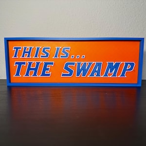 May include: A rectangular sign with a blue frame and an orange background. The sign reads "THIS IS... THE SWAMP" in blue, outlined white, block letters. The sign is resting on a dark surface.