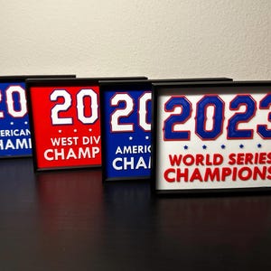 May include: Four framed signs with the number "20" and "2023" in white and blue, with text indicating "American Champs", "West Div Champ", "American Champs", and "World Series Champions". The frames are black.