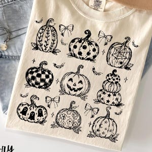 May include: An ivory short-sleeve t-shirt featuring a black ink graphic print of various pumpkins, bows, bats, and stars. The pumpkins display diverse patterns including stripes, polka dots, spiderwebs, checkers, ghosts, and floral motifs.