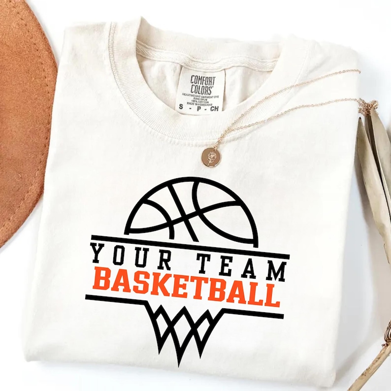 May include: Cream-coloured t-shirt with a basketball graphic. The design features a black basketball, the words "YOUR TEAM" in black, and "BASKETBALL" in orange. A gold necklace with a pendant is draped on the shirt.