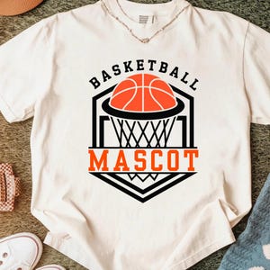 May include: White t-shirt with a basketball graphic. The design features a basketball, net, and the words "BASKETBALL MASCOT" in black and orange. The shirt is made of a soft material.