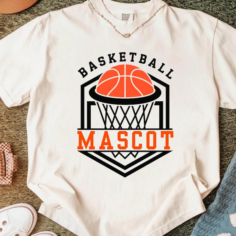 May include: White t-shirt with a basketball graphic. The design features a basketball, net, and the words "BASKETBALL MASCOT" in black and orange. The shirt is made of a soft material.