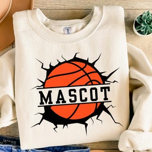 May include: Cream-coloured sweatshirt with a basketball graphic breaking through a cracked black background. The word "MASCOT" is displayed in bold, black capital letters across the basketball. The sweatshirt is made of a soft material.