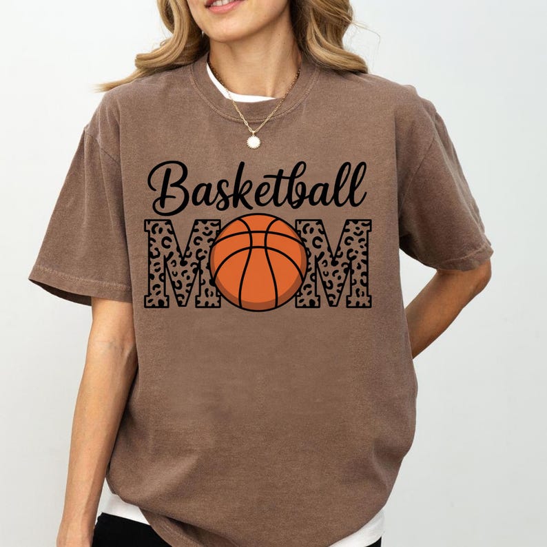 May include: A brown t-shirt with the words "Basketball MOM" in black script and leopard print. An orange basketball is in the center of the word "MOM". The shirt has short sleeves and a crew neck.
