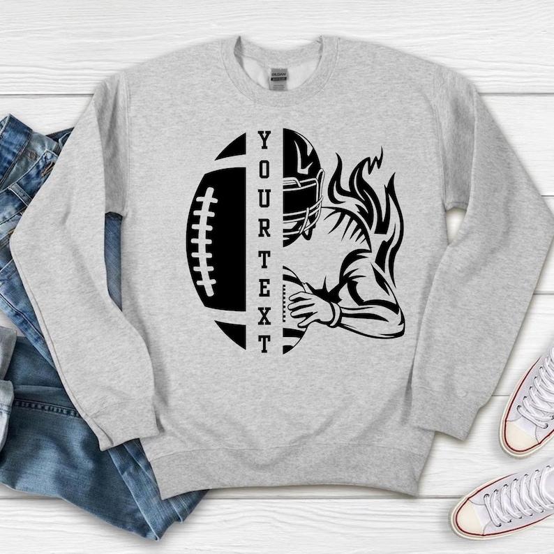 May include: A light gray sweatshirt featuring a black graphic of a football and a football player. The football is split in half with the text "YOUR TEXT" vertically. The player is holding the ball with flames rising behind them.