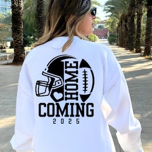 Könnte beinhalten: Weißes Sweatshirt mit einem schwarzen Grafikdesign mit einem Footballhelm, Football und den Worten "HOME COMING 2025". Das Design ist auf der Rückseite des Sweatshirts zentriert.