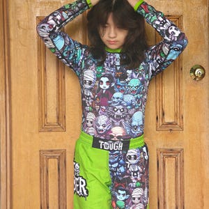 Könnte beinhalten: Ein langärmeliges Shirt und Shorts-Set mit einem farbenfrohen Monster-Motiv. Das Shirt und die Shorts zeigen eine Vielzahl von Cartoon-Monstern in Blau-, Grün-, Lila- und Weißtönen. Die Shorts haben das Wort "TOUGH" auf dem Bund.