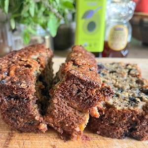 May include: A loaf of baked bread sliced into three distinct pieces, featuring a dark brown, textured crust and a dense interior crumb speckled with dark berries. The bread displays a rustic, uneven surface with visible grains and fruit throughout.
