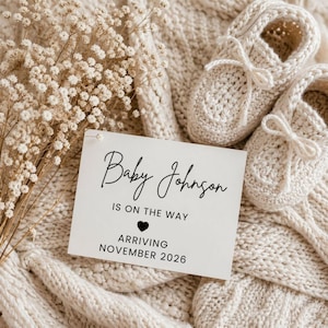 May include: A cream-coloured baby announcement card with the text "Baby Johnson IS ON THE WAY ARRIVING NOVEMBER 2026" rests on a knitted blanket. Two small, cream-coloured knitted baby booties are next to the card. Dried flowers are in the background.