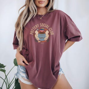 May include: A maroon-colored t-shirt with the words "POTTERY SOCIAL CLUB" in a circular design. The graphic in the center depicts a pottery vessel held by two hands. The word "NOEL" is below the graphic.