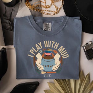 May include: A blue Comfort Colors t-shirt with the text "I PLAY WITH MUD" in a circular design. The design includes a pottery wheel, hands, and a paintbrush. The shirt is a size XL-TG.
