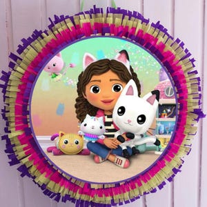 Könnte beinhalten: Eine runde Piñata mit einem farbenfrohen Design. Das zentrale Bild zeigt eine Zeichentrickfigur mit Katzenohren, die zwei Plüschtiere mit Katzenmotiven hält. Die Piñata hat Schichten aus lila, rosa und gelben Fransen und ein grünes Band zum Aufhängen.