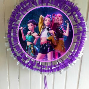 Op de afbeelding: Een ronde piñata met een centrale afbeelding van drie geanimeerde vrouwen die microfoons vasthouden. De piñata is versierd met paarse en zilveren franjes. De afbeelding is omgeven door een roze rand.