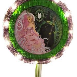 May include: Round piñata with a green and silver fringe border, featuring a pink and green image of characters from the musical "Wicked". The piñata has a white and green ribbon hanging from the bottom and is decorated with small lights.