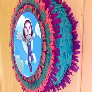 Könnte beinhalten: Eine bunte Piñata in Kreisform mit einem Bild eines Comic-Superhelden in einem weißen Anzug mit einem Spinnen-Symbol auf der Brust. Die Piñata ist mit rosa, türkisfarbenem und violettem Seidenpapier verziert.