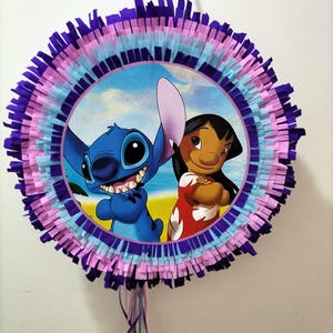 Może przedstawiać: Okrągła niebiesko-fioletowa piñata z obrazkiem Stitch'a i Lilo z filmu Disneya Lilo & Stitch. Piñata jest ozdobiona niebieskimi, różowymi i fioletowymi wstążkami z papieru.