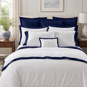 May include: A bedding set featuring a white duvet cover and matching pillowcases with a solid navy blue border. The bed is layered with navy blue quilted shams and a small white rectangular accent pillow centered in the front of the arrangement.