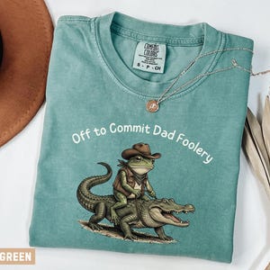 May include: Light green Comfort Colors t-shirt with the phrase "Off to Commit Dad Foolery" in white. The shirt features a cartoon frog wearing a cowboy hat and vest riding an alligator. A gold necklace is draped on the shirt.