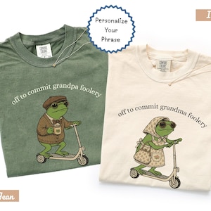 May include: Two crew neck t-shirts in blue jean and ivory colors. The blue jean shirt features a frog in a brown sweater and cap riding a scooter with the text off to commit grandpa foolery. The ivory shirt shows a frog in a floral apron and headscarf riding a scooter with the text off to commit grandma foolery.