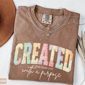 May include: Espresso-colored t-shirt with the word "CREATED" in colorful, patchwork-style letters. Below, the text reads "Ephesians 2:10 with a purpose." A silver necklace with a small pendant rests on the shirt. A brown hat and dried plant are in the background.