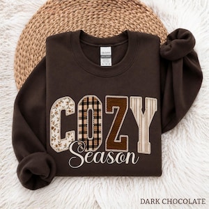 May include: A dark chocolate brown crewneck sweatshirt featuring the word COZY in large, fabric-appliqued letters with varied patterns including floral, plaid, and solid textures, with the word Season written in white cursive script below.