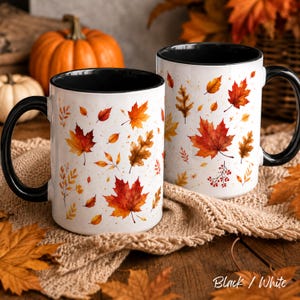 May include: Two white ceramic mugs with black handles and interiors, featuring a repeating pattern of colorful autumn leaves in shades of red, orange, and yellow, with small brown speckles scattered across the surface of the mugs.