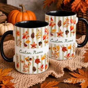 May include: Two ceramic mugs with black handles and interiors, featuring a pattern of autumn leaves, pinecones, and acorns over vertical sage green stripes. Each mug has a white horizontal band with the text Custom Name printed in green.