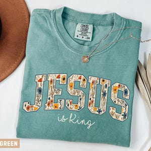 May include: A light green short-sleeve t-shirt featuring the word JESUS in large block letters filled with a floral pattern, with the text is King written below in white script. The shirt has a Comfort Colors label and a crew neckline.
