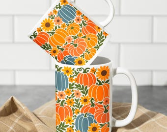May include: Two white ceramic mugs with a fall-themed design. The mugs feature a pattern of orange pumpkins, blue pumpkins, yellow sunflowers, and green leaves. The mugs have large white handles and are stacked on top of each other.