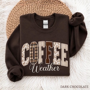 May include: A dark chocolate brown crewneck sweatshirt featuring the word COFFEE in large, block-style letters with varied patterns including plaid, floral, and solid brown, with the word Weather written below in a white cursive script font.