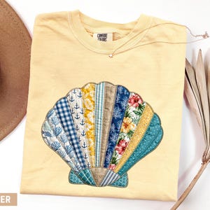 May include: Butter-colored t-shirt featuring a seashell design. The seashell is made of various fabric patterns, including blue anchors, floral prints, and stripes. A gold necklace is draped across the shirt.