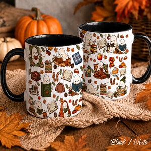 May include: Two white ceramic mugs with black handles and interiors, featuring a repeating pattern of cartoon ghosts reading books, pumpkins, autumn leaves, plaid blankets, candles, and slices of pie on a white background.