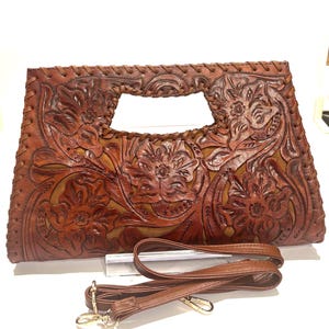 May include: A brown leather clutch with a floral embossed design. The bag has a rectangular shape with a handle cutout and a detachable shoulder strap. The leather is a warm, reddish-brown color.