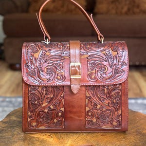 Puede incluir: Un bolso de mano de cuero marrón con intrincados grabados florales y de volutas. El bolso presenta un asa superior, un cierre de solapa con hebilla y una correa de hombro desmontable. El cuero tiene un tono cálido y rico.