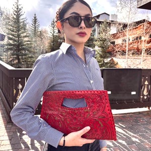 Puede incluir: Un bolso de mano de cuero rojo con un diseño floral en relieve. El bolso tiene forma rectangular con un asa recortada. El fondo incluye una persona con gafas de sol y una camisa a rayas.