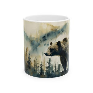 May include: White ceramic mug featuring a watercolor painting of a grizzly bear in a forest setting. The design includes a bear, trees, and mountains in shades of brown, blue, and beige. A nature-themed coffee mug.