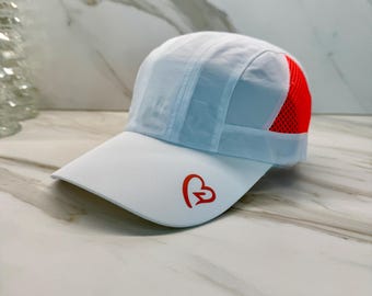 Könnte beinhalten: Hellblaue Baseballkappe mit gebogenem Schirm. Die Kappe hat ein rotes herzförmiges Logo auf der Vorderseite und ein rotes Mesh-Panel an der Seite. Die Kappe ist aus leichtem Material gefertigt.