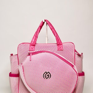 Puede incluir: Un bolso tote de malla rosa con una bolsa con cremallera a juego. El bolso tiene un asa superior y bolsillos laterales. La bolsa y el bolso presentan un logotipo negro. El bolso está diseñado con un patrón rosa y blanco.