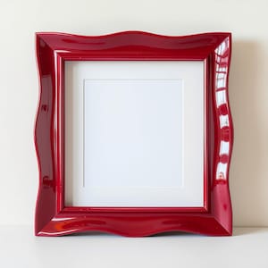 May include: A glossy red picture frame with a wavy, ornate design. The frame has a white matting around the central opening. The frame is square and suitable for displaying photos or artwork.