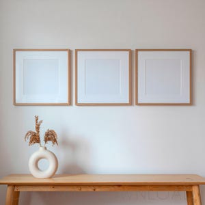 May include: Three square wooden picture frames with white matting are mounted on a wall above a light wood console table. A round, off-white ceramic vase containing dried beige grasses sits on the left side of the table surface.