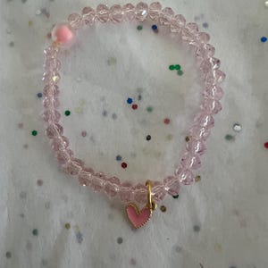 Può includere: Un braccialetto di perline rosa con un ciondolo a forma di cuore dorato. Il braccialetto è fatto di piccole perline sfaccettate e presenta un piccolo pompon rosa. Il ciondolo a forma di cuore è rosa con un bordo dorato.