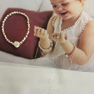 Puede incluir: Una pulsera de cuentas plateadas con una perla grande y redonda. La pulsera se muestra sobre un cojín burdeos. La imagen también muestra a un niño con pulseras y anillos.