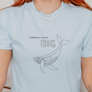 May include: Light blue t-shirt with the text "God loves you so BIG" and a line drawing of a whale. The shirt is a soft, casual style, suitable for everyday wear.